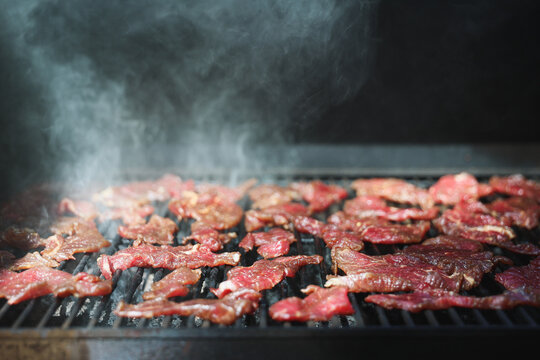 Slices of round steak rest on a pellet grill smoker, their glossy surface glistening in light as smoke rises, capturing the slow process of beef jerky preparation.