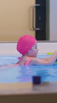 Young girl in pink swim cap and suit floats on stomach. Yellow rubber duck appears near wall with blue objects. Colored objects such as green kickboard or small white toy