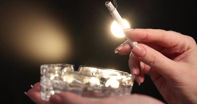 Woman flicking ash from cigarette into ashtray on black background, closeup. Camera moving forward