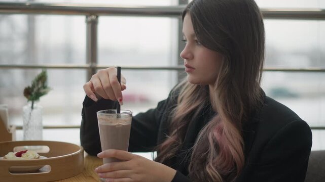 Young woman sits at round cafe table on urban terrace, stirring tall glass of iced coffee with metal spoon while gazing slightly upward; gentle wrist movement and relaxed posture create quiet