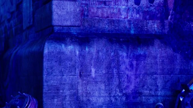 A stone wall stands prominently in an urban area. Blue light reflects on its surface, creating striking visual effects. People walk by, unaware of the artful display at night.