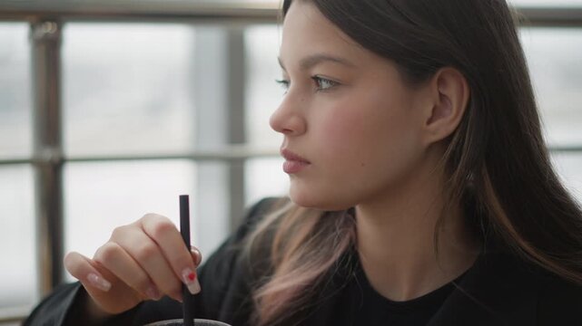 Young woman seated indoors lifts straw to lips and sips iced drink while looking outward with thoughtful calm; soft window light, shallow focus, and quiet lifestyle atmosphere shape scene