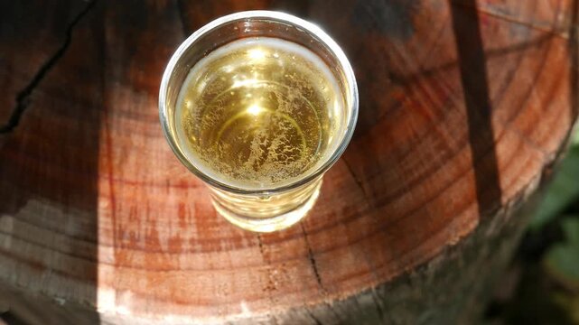 Close-up of a glass of beer on a table in the sun. A zoom-out reveals a small glass of draft beer on a wooden table. A glass of ice-cold beer under random rays of sunlight. Drinking beer, POV.