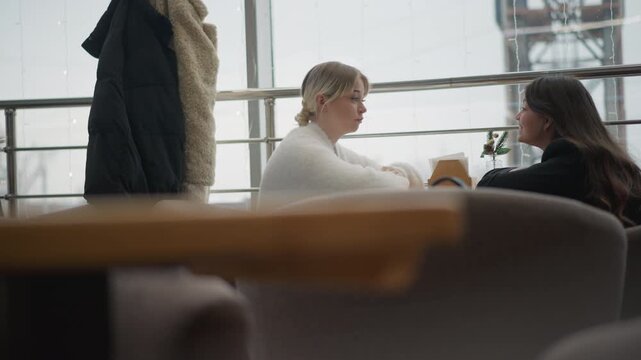 Two women sit facing each other across table in bright cafe, one leaning forward with hands near tabletop while both share calm attentive expressions and relaxed conversation, with women