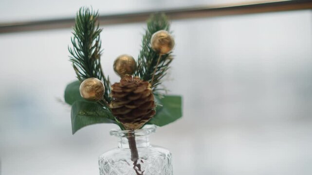 Close-up of seasonal tabletop arrangement showing central pinecone with evergreen sprigs, glossy holly-like leaves, and three small gold spherical ornaments placed in textured clear glass vase with