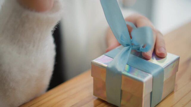 Close-up eye-level view of child's hands untying pastel blue ribbon from small iridescent gift box on wooden table