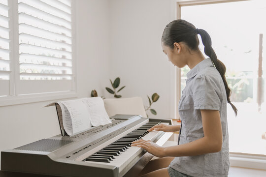 piano, music, teen, home, playing, keyboard, practicing - A young mixed Asian teen focuses on practicing a song on her digital piano in a bright living room, capturing a peaceful moment.