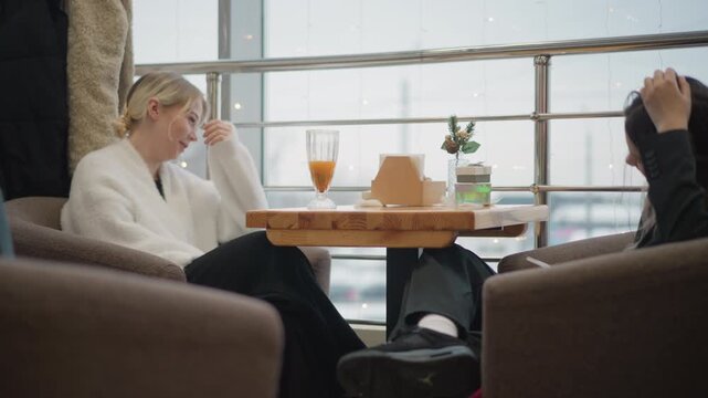 Two women sit across round wooden table near large window in cozy cafe, sharing relaxed conversation with smiles, hand gestures, casual posture, drinks, takeaway box, and soft daylight filling warm