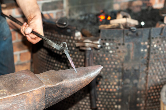 A forger wearing blue jeans and a T-shirt holds fire tongs and a twisted metal bolt on a vintage anvil. There's a rounding hammer with a wooden handle in the blacksmith's foundry, near the furnace.