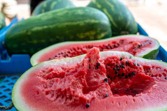 Fresh, elongated watermelon with a transverse cut. The pink, chunky inside has lots of black seeds. The outside of the fruit has a striped green and yellow rind. The watermelon is in a blue tray. 