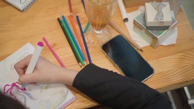 Overhead view shows hand sketching and erasing inside spiral sketchbook on wooden desk, with colored pencils, cup, phone case, stationery, and scattered tools suggesting active creative work