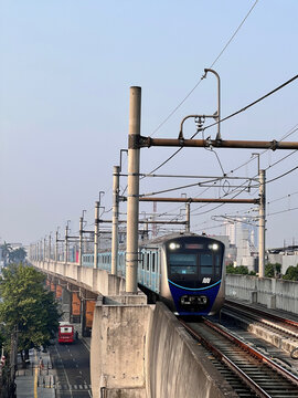 A modern electric commuter train traveling along an elevated railway in a busy city environment, approaching on parallel tracks beneath a network of overhead electrical wires and supporting poles.