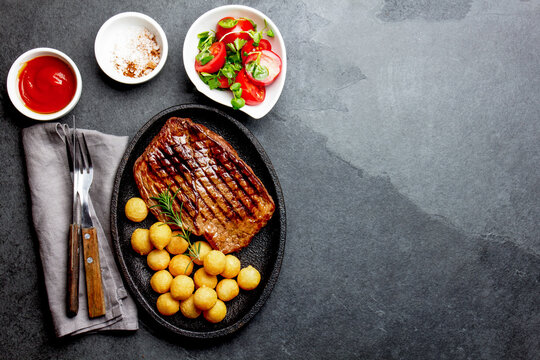 Grilled beef steak served on cast iron plate with tomato salad and potatoes balls. Barbecue, bbq meat beef tenderloin. Top view, slate background