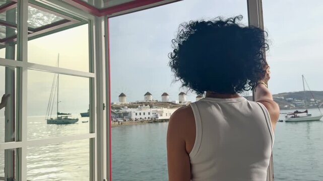 Woman Enjoys Mykonos Sunset View from Window Overlooking Aegean Sea