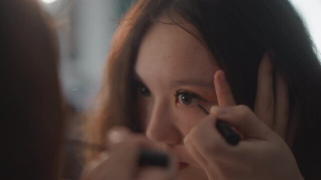 Intimate beauty scene showing young woman having eyeliner applied to upper lash line while hands gently steady eyelid, with focused upward gaze, soft warm lighting, and professional care