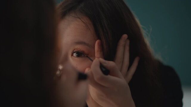Close-up of young woman applying eyeliner at outer eye while gazing into mirror, hand holding brush as fingers lift eyelid for precise delicate makeup motion in soft indoor light, with woman