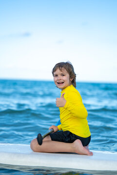 Child on a paddleboard on summer sea. Child with paddle near sea skyline. Smiling kid learns summer water sports. Child gives thumbs-up beside ocean waves.