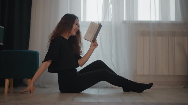 Young woman sits on pale wooden floor beside bright window, holding book at eye level while pages flutter softly and hair moves slightly in calm, cozy home interior, with woman, reading, sitting