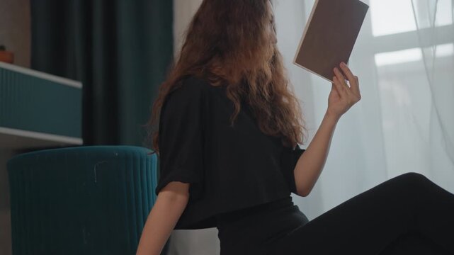 Cropped view of woman on hardwood floor with legs extended, torso leaning back and one hand braced for support while other holds book near face, creating calm indoor reading moment