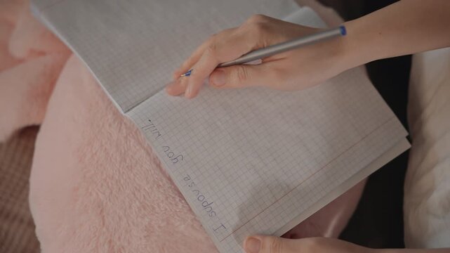 Overhead close-up shows hands writing with pen in open lined notebook on textured pink blanket beside soft pillow, revealing calm home journaling, focused note making, and gentle motion