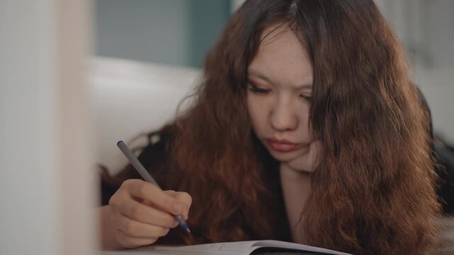 Close view of young woman with curly brown hair leaning over notebook while helper holds spoon near mouth, showing focused multitasking, gentle support, warm light and quiet domestic scene
