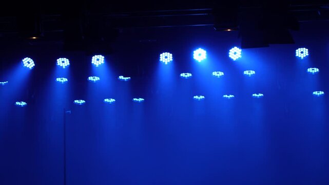 Horizontal wide shot of a stage lighting rig with multiple LED lights emitting deep blue volumetric beams downwards through smoke in a dark venue