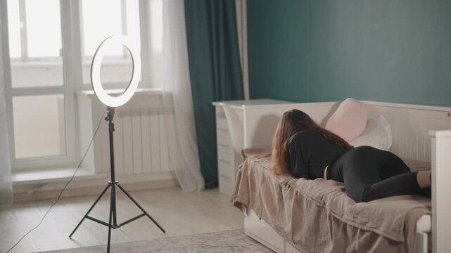 Young woman lies on side on sofa with head on pillows and knees slightly bent, facing away from camera in softly lit home interior, while ring light on tripod stands nearby and adds gentle glow
