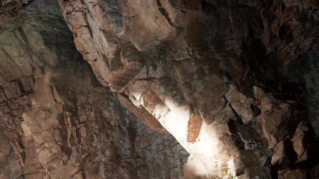Bright light shines on the surface of a large rock formation inside a cave. Textured stone walls surround the area. The setting shows natural geological features and shadows.