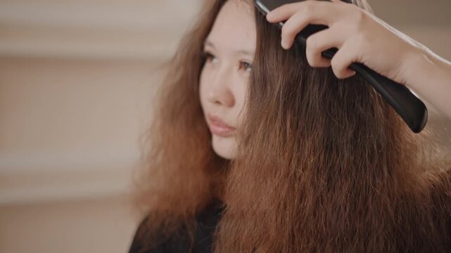 Medium close-up of person combing long, frizzy red-brown hair with paddle brush; visible downward stroke smooths textured, voluminous strands while flyaways and split ends separate as brush passes