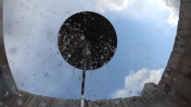 Water droplets falling from a large round shower head, framed by a wooden enclosure and bright summer sky