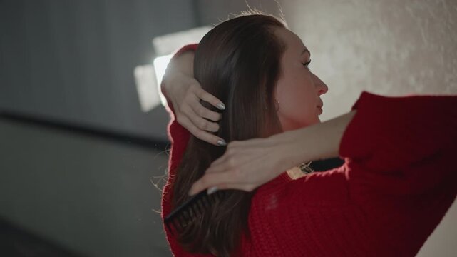 Side-profile view of woman wearing red sweater combing long brown hair with raised hand, showing subtle movement, soft backlight glow, and gentle grooming action in indoor portrait setting