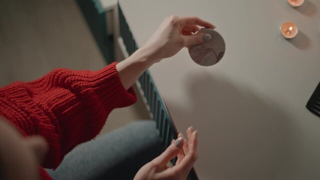 Top-down view of adult hands in red knit sweater opening, closing, and placing round compact mirror with silver case on light tabletop beside lit tealight candles and comb, creating cozy beauty scene