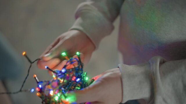 Close slightly elevated view of hands untangling and spreading compact bundle of glowing multicolored LED string lights, with visible finger movement, soft indoor light, cozy mood and festive decor