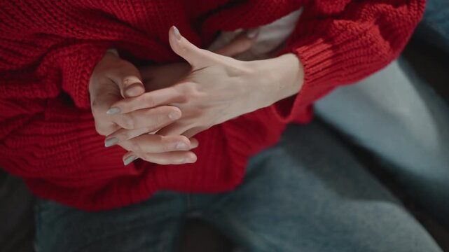 Overhead close-up showing adult hands clasped together and gently rubbing over lap while seated indoors, with red knit sweater, denim pants, soft light, and subtle fidgeting detail