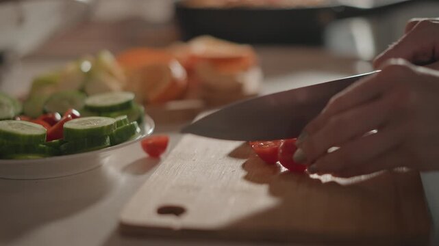 Close-up kitchen scene showing hands slicing cherry tomatoes on wooden cutting board beside plated cucumber salad, with visible juices, seeds, knife movement and fresh meal preparation
