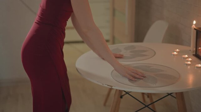 Young woman in red dress moves around round dining table, placing and adjusting placemats and plates with both hands while lit tealight candles and decorative lantern create warm soft side lighting