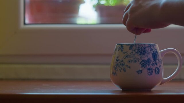 Coffee cup hand stirring spoon inside a patterned ceramic mug on a wooden windowsill. Morning routine breakfast beverage, hot tea preparation with blurred window background, static shot.