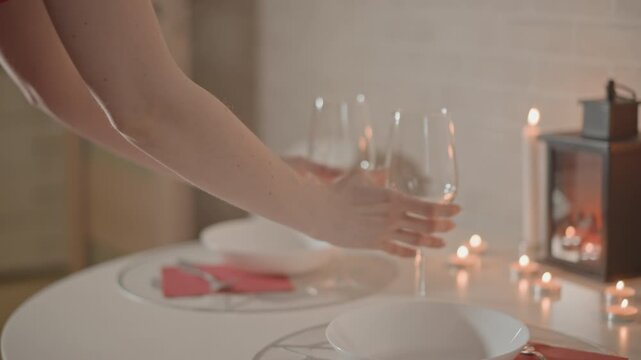 Two empty wine glasses are gently set together on round white table next to white plates and folded red napkins while small lantern and tealight candles cast warm highlights and soft reflections on