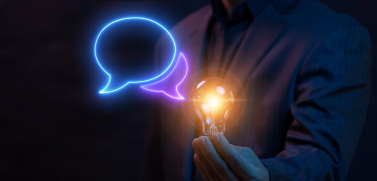 Businessman holding bright glowing lightbulb in dark with glowing double chat speech bubbles frame beside display. Creative brainstorming dialogue, customer feedback idea, and communication concept.