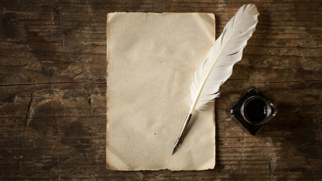 Old parchment paper with white feather quill and black inkwell resting on rustic wooden desk surface