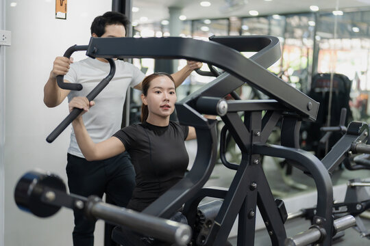 Asian woman working out on chest press machine with male personal trainer assistance in gym