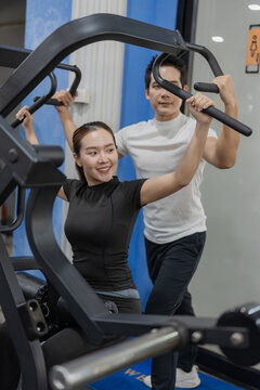 Asian woman working out on chest press machine with male personal trainer assistance in gym
