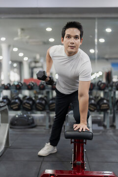 Asian man sitting on workout bench doing bicep curls with dumbbells in gym