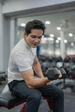 Asian man sitting on workout bench doing bicep curls with dumbbells in gym