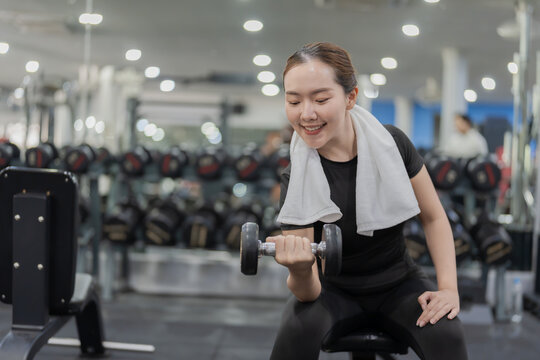 Smiling Asian woman sitting on workout bench doing bicep curls with dumbbell in gym