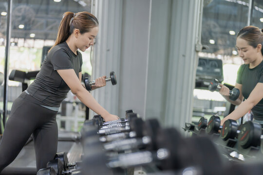 Asian woman taking dumbbell from rack in front of mirror in gym