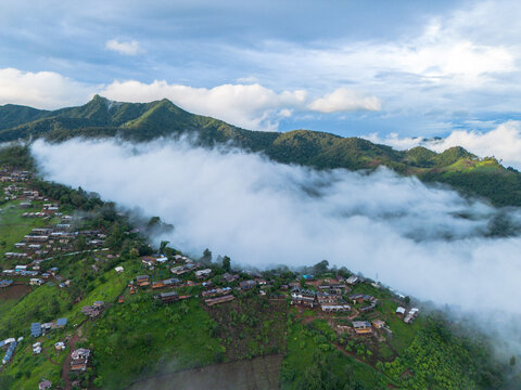 Mountain village covered by rolling cloud in lush green highland landscape, with scattered homes and farmland beneath dramatic blue sky and layered forested ridges