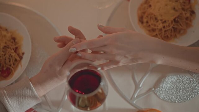 Top-down view showing two adults holding hands across white tablecloth in warm restaurant setting, with pasta, wine, plates, cutlery, and soft light creating intimate evening mood, with hand, holding