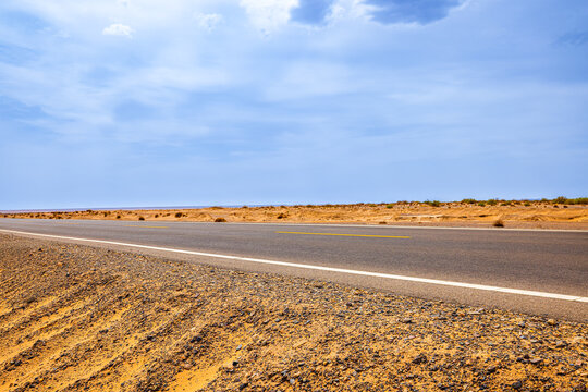 Empty paved highway crosses the arid Dahaidao desert in Xinjiang, China, offering travel, transportation, and expansive landscape imagery under a cloudy blue sky.
