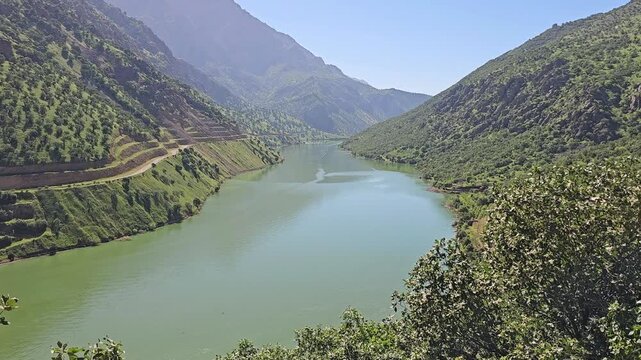 Tranquil blue river winds beneath a lush green hillside, bordered by a dirt path and dense trees under a bright spring sky.
📍 Hawraman Takht, Kurdistan, Iran 